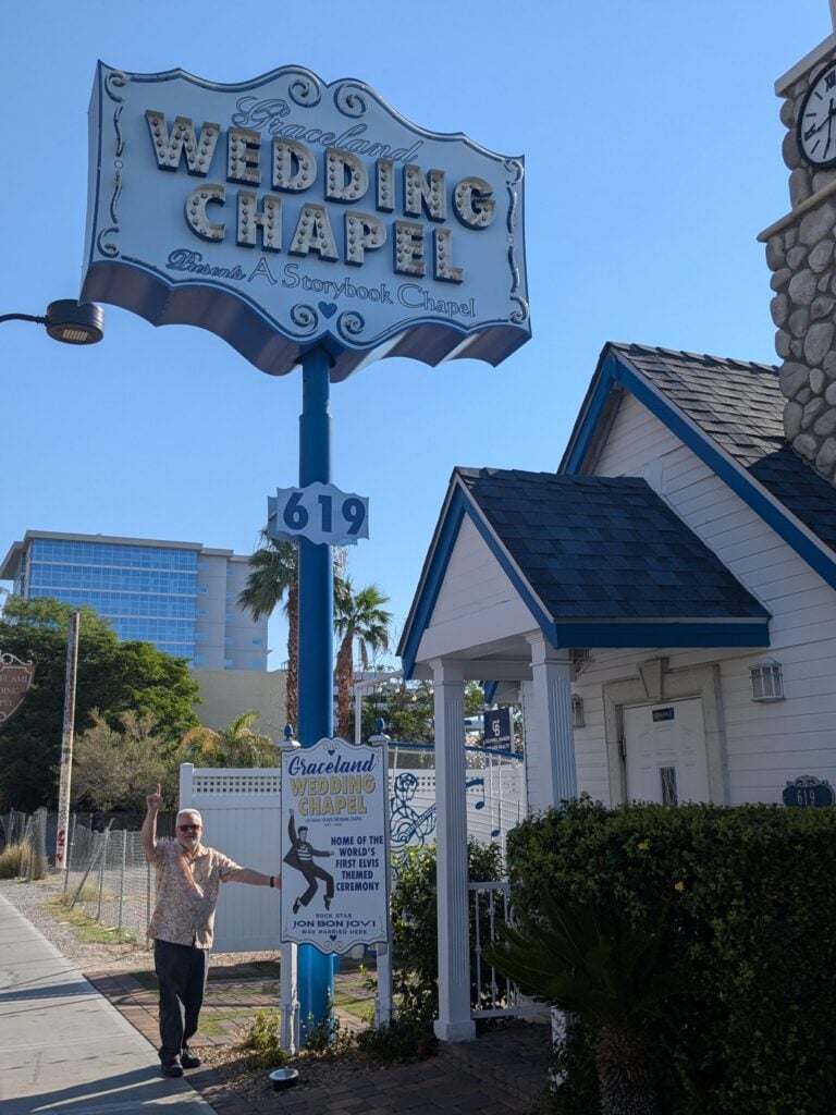 A man stands smiling and waving outside the white and blue Graceland Wedding Chapel beneath a large vintage sign on a sunny day. The chapel is surrounded by palm trees and other buildings.