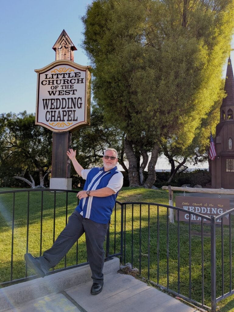 A smiling man in sunglasses and a blue shirt poses with outstretched arms in front of a large Little Church of the West Wedding Chapel sign, surrounded by green grass and trees on a sunny day.