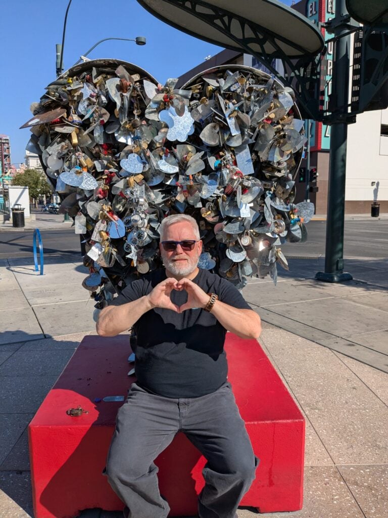 A man with gray hair and a beard, wearing sunglasses and a black shirt, sits on a red platform in front of a large heart-shaped sculpture covered in locks, making a heart shape with his hands.