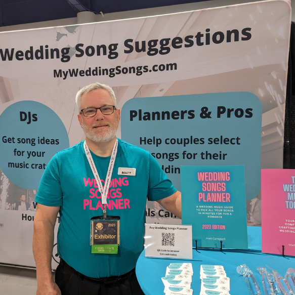 A man wearing a teal Wedding Songs Planner shirt and exhibitor badge stands at a booth with displays suggesting wedding songs. The booth backdrop reads Wedding Song Suggestions and promo materials are on the table.