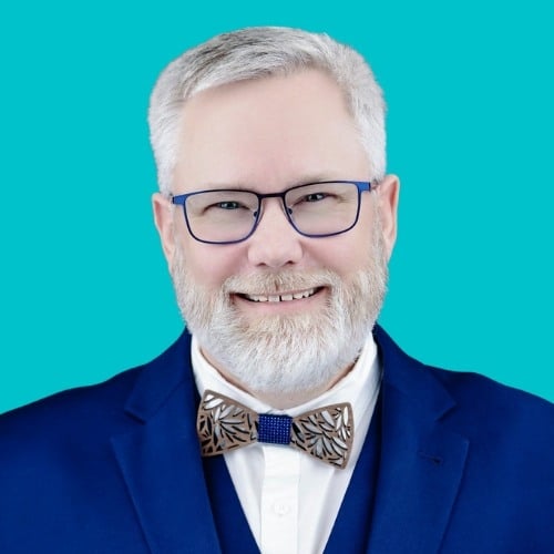 A smiling man with gray hair and beard wearing glasses, a blue suit, white shirt, and a decorative wooden bow tie, posing in front of a solid turquoise background.